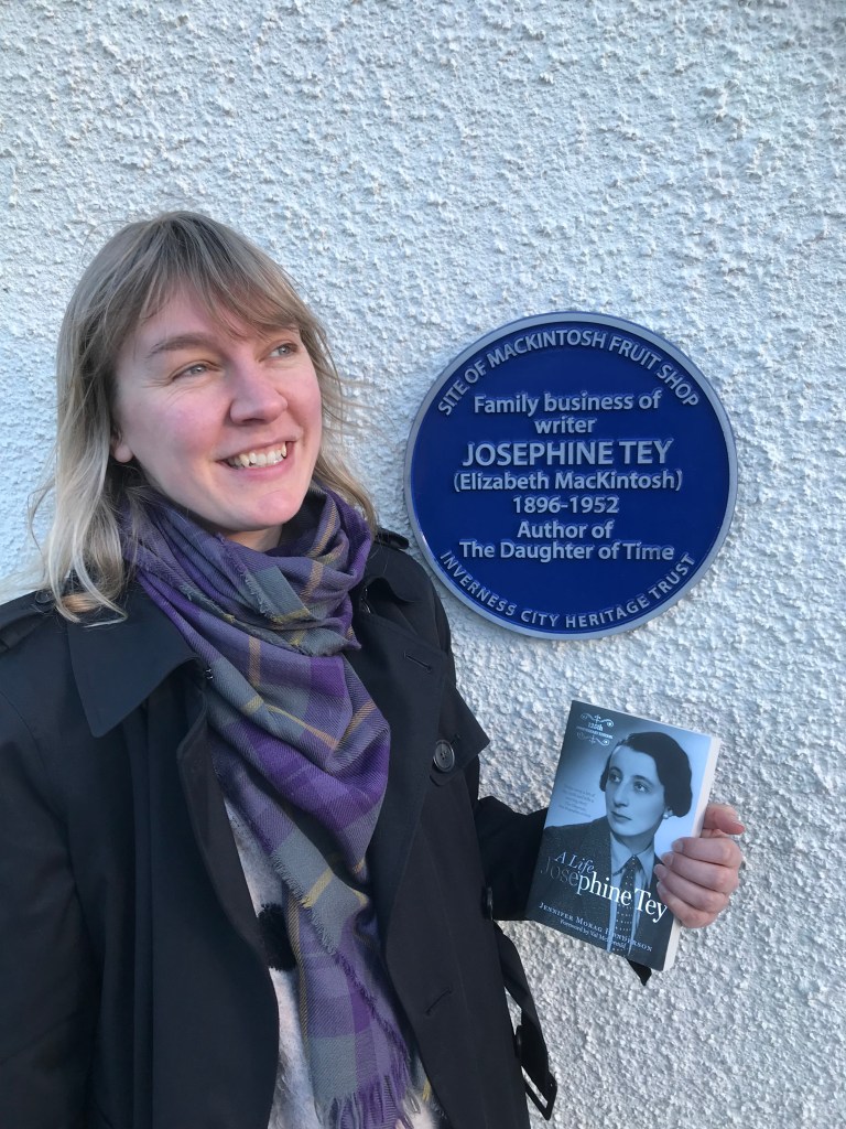 Jennifer Morag Henderson standing in front of the new blue plaque for Josephine Tey in Inverness, holding her biography of Tey.